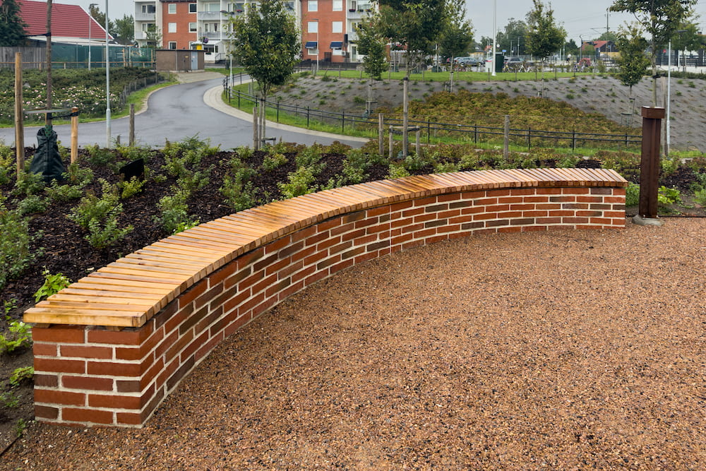 Svängd murad bänk i tegel med plantering och gångväg i bakgrunden.